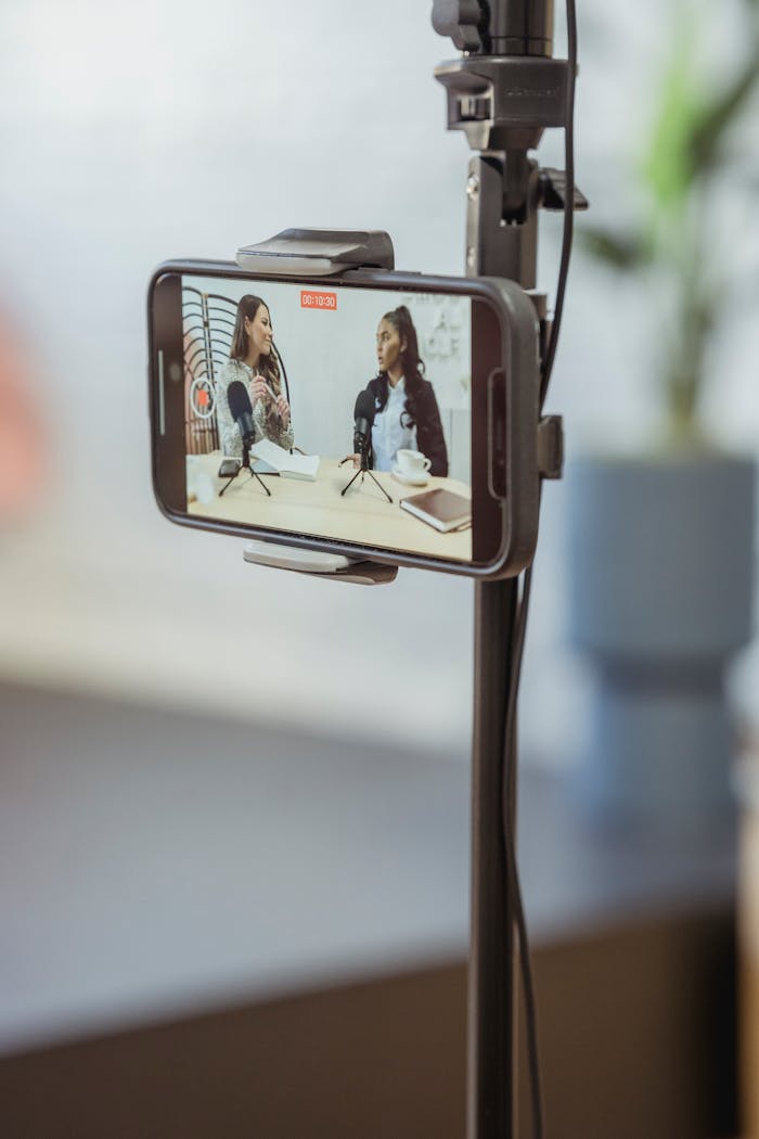 why-choose-us-02 Young diverse women sitting at table with microphones and gadgets and talking while recording podcast on smartphone placed on tripod in studio